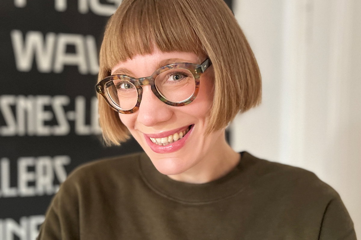 Porträt einer Person mit kurzem, geradem Haarschnitt und runder Brille vor unscharfem Innenhintergrund. Die Person trägt ein dunkles, schlichtes Oberteil. Im Hintergrund sind verschwommene weiße Schriftzüge auf dunkler Fläche zu erkennen.