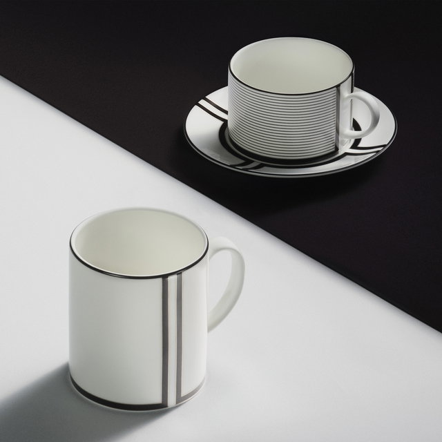 White and black table top with a white mug featuring black edging sat on the white tablecloth, and a white and black patterned teacup & saucer displayed on the black cloth.