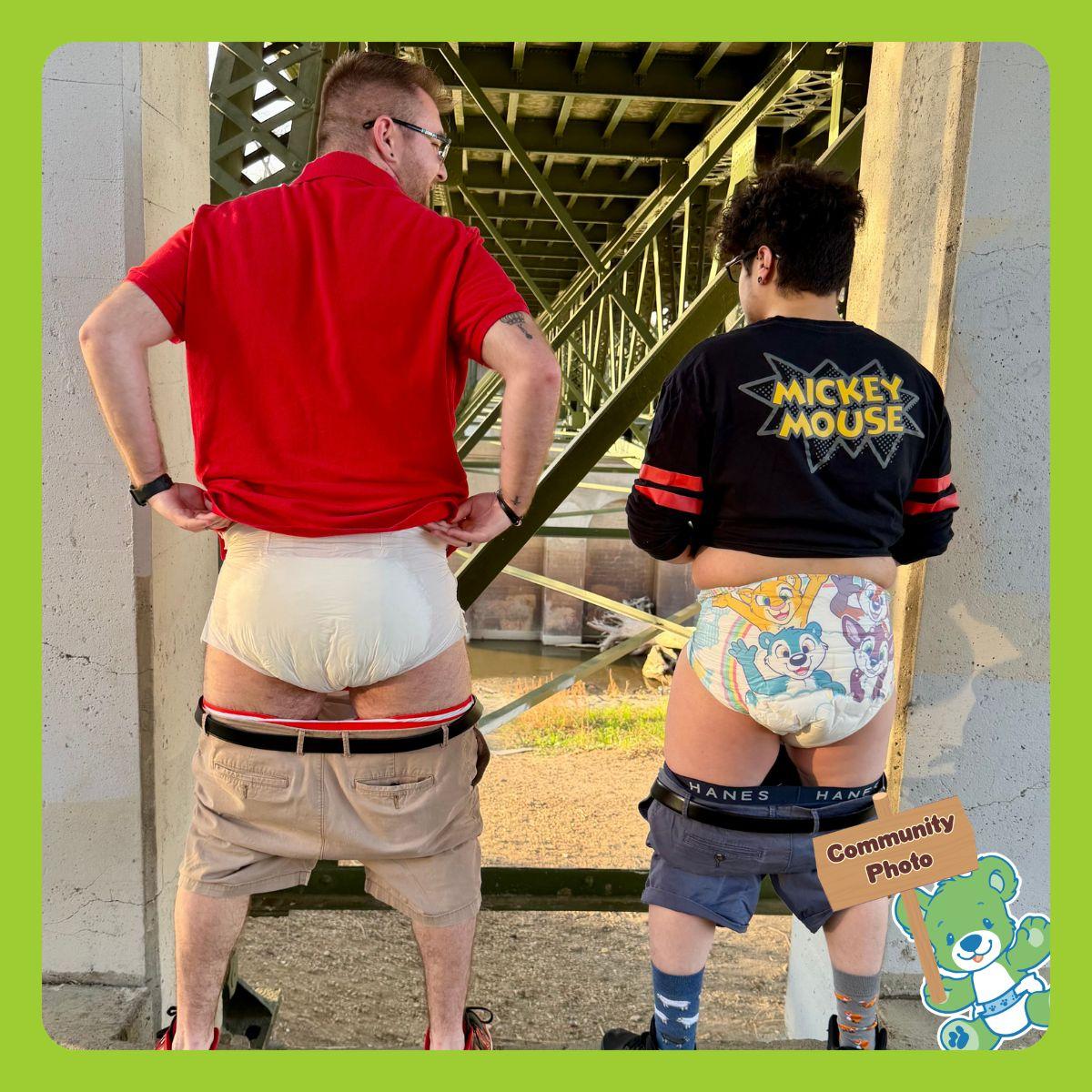 Image of two human pup players, Pup Ashton (on the left) and Pup Cryptic (on the right). They are standing under a bridge with their pants down showing Ashton is wearing a PeekABU adult diaper, while Cryptic is wearing a TinyTails adult diaper. Both of the pups are facing away from the camera showing their backs,