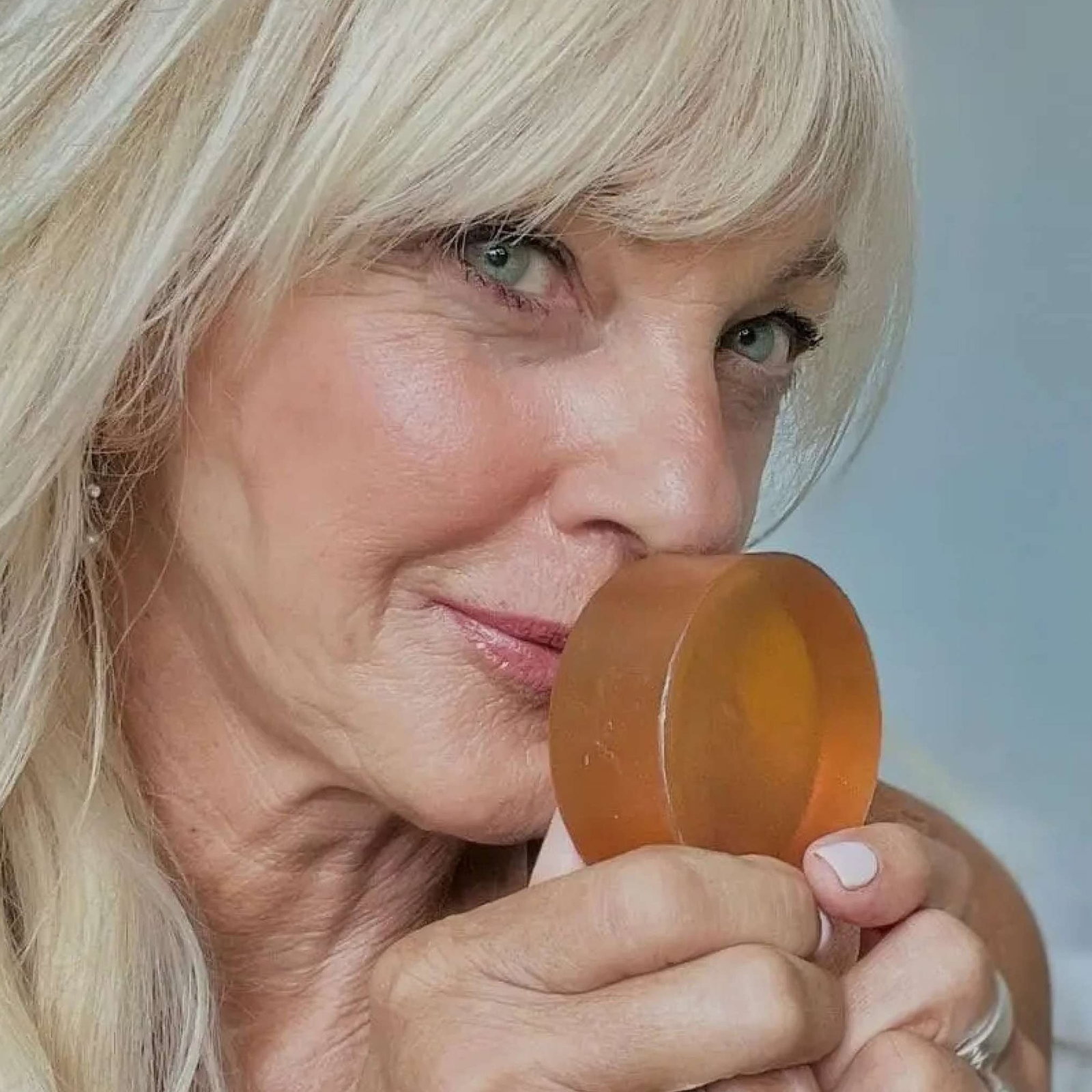 A close-up shot of a mature woman with blonde hair, her face tilted as she brings a translucent, amber-colored soap bar close to her nose. Her light blue eyes are looking forward, and her skin shows signs of age. (square)