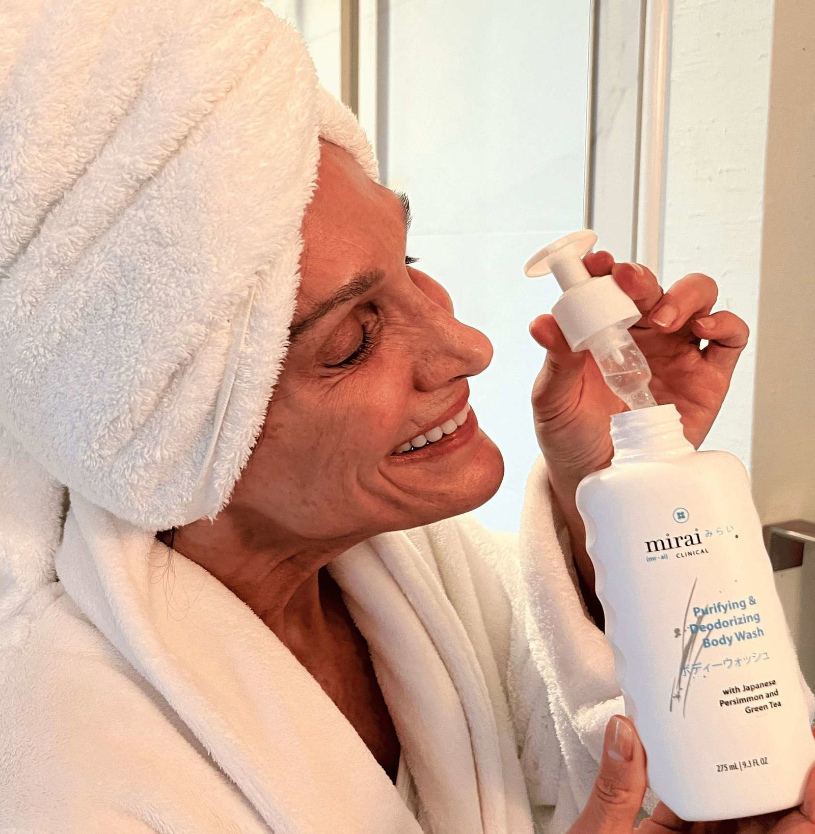 A woman wearing a white towel wrap on her head and a white bathrobe is holding a bottle of Mirai Clinical Purifying & Deodorizing Body Wash and pressing the pump. (square)