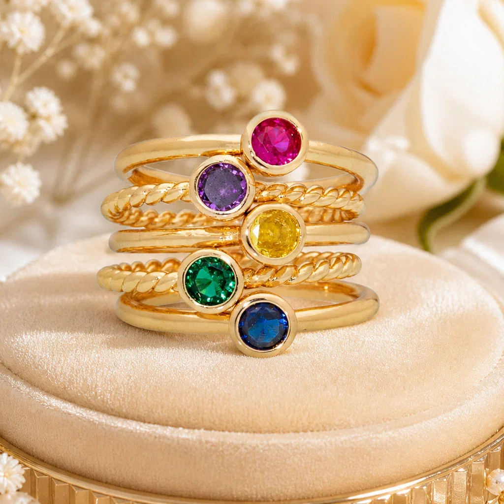 Five stacked gold rings with colorful gemstones: ruby, amethyst, citrine, emerald, and sapphire on a beige display stand.