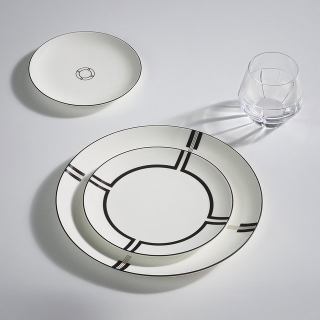 New Vera Wang Luxe Graphite tableware pieces latered and presented onn a white tablecloth with a crystal tumbler. A side plate sits at the top left, with the crystal tumbler at the top right, followed by a small plate layered on top of a dinner plate in the bottom middle.