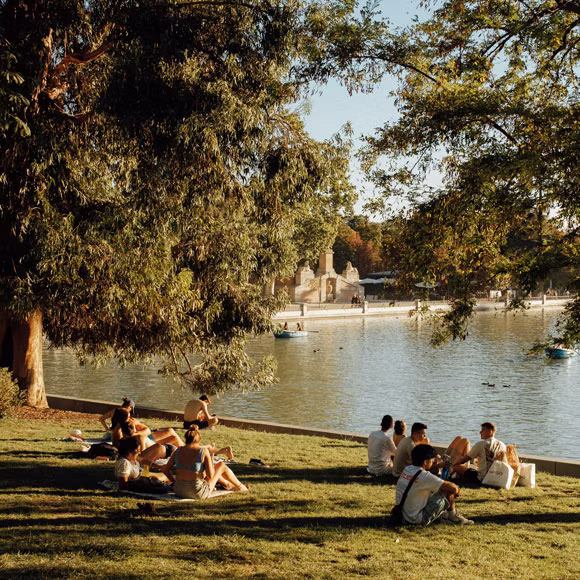 People sitting on the grass along the water’s edge in Madrid’s Retiro Park, linking to BA High Life site.