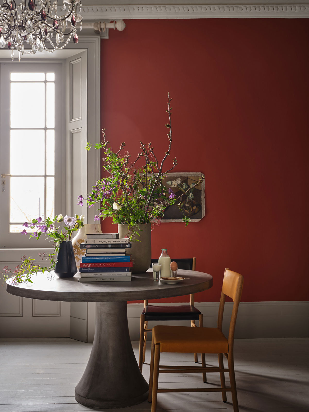 A dramatic dining space with bold red walls, where a round table, simple wooden chairs and overflowing garden flowers create a welcoming scene. With an abundance of natural light coming from a nearby window, the space feels warm and expressive, the perfect place to catch up with friends.