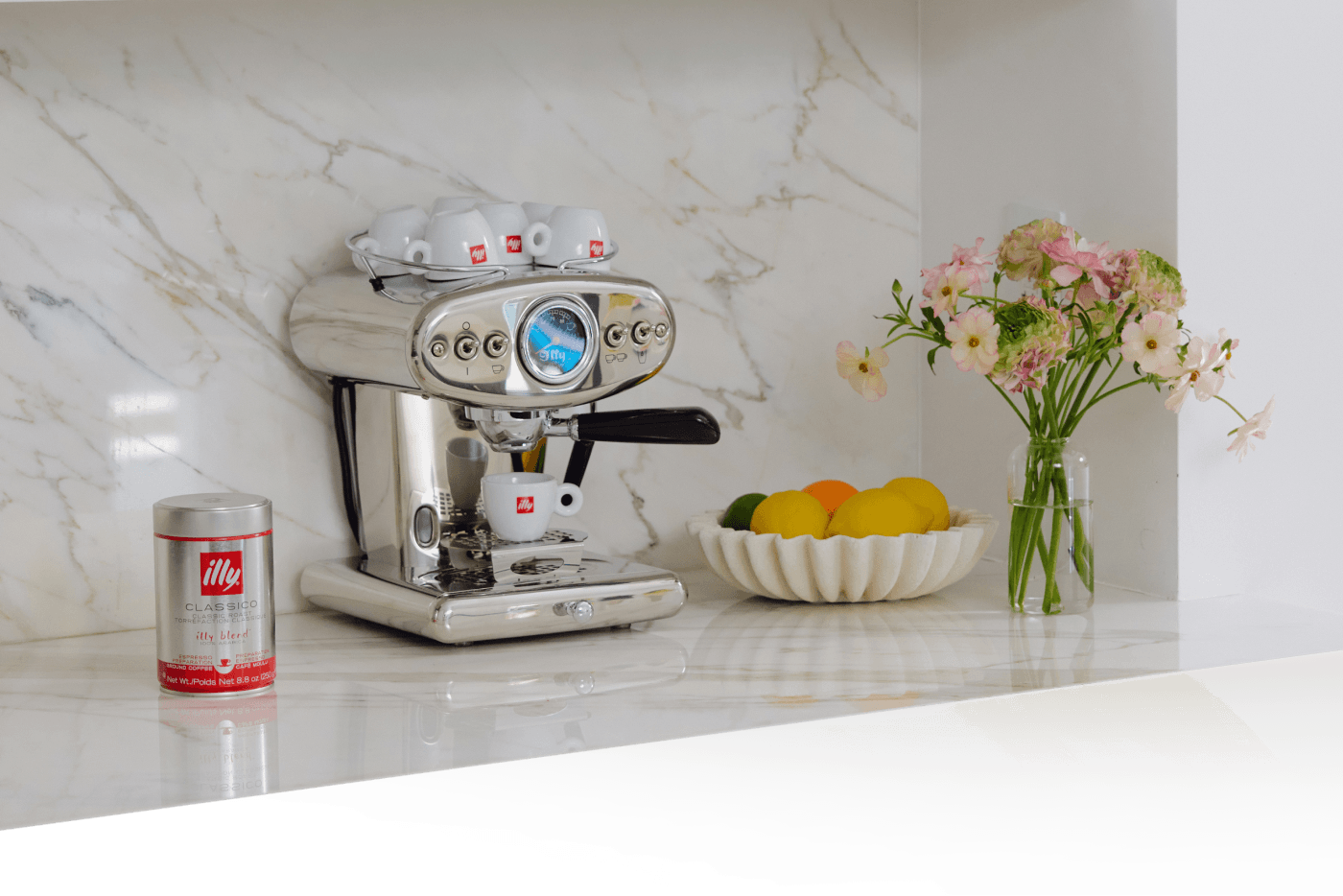 Stylish kitchen counter with an espresso machine, illy coffee tin, fresh flowers, and a bowl of citrus fruit