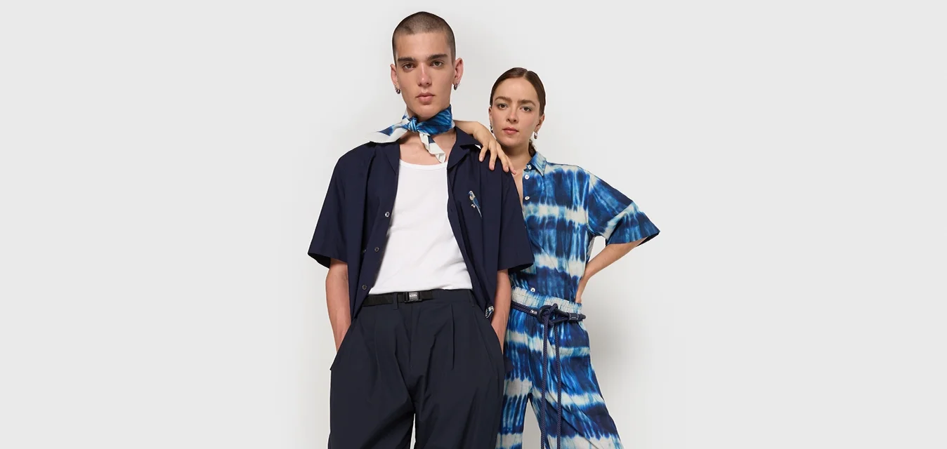 Twee modellen staan samen. De één draagt een marineblauw shirt en wijde broek; de ander heeft een blauw tie-dye jumpsuit aan. Beide hebben minimale accessoires.