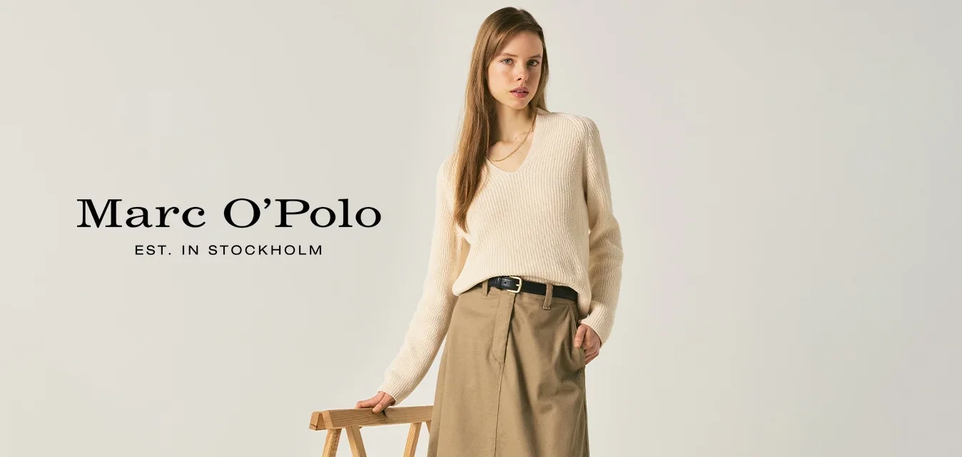 Vrouw draagt een crèmekleurige trui, beige midi-rok, zwarte riem en zwarte loafers, staat naast een houten inklapbare standaard tegen een effen achtergrond.