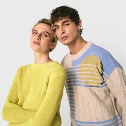 Dos jóvenes adultos, uno con un suéter amarillo y otro con un suéter de patrón beige y azul, de pie juntos contra un fondo blanco.
