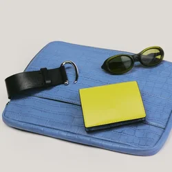 Bolso de mano azul texturizado con cinturón de cuero negro, cartera amarilla y gafas de sol ovaladas oscuras dispuestos sobre un fondo claro.