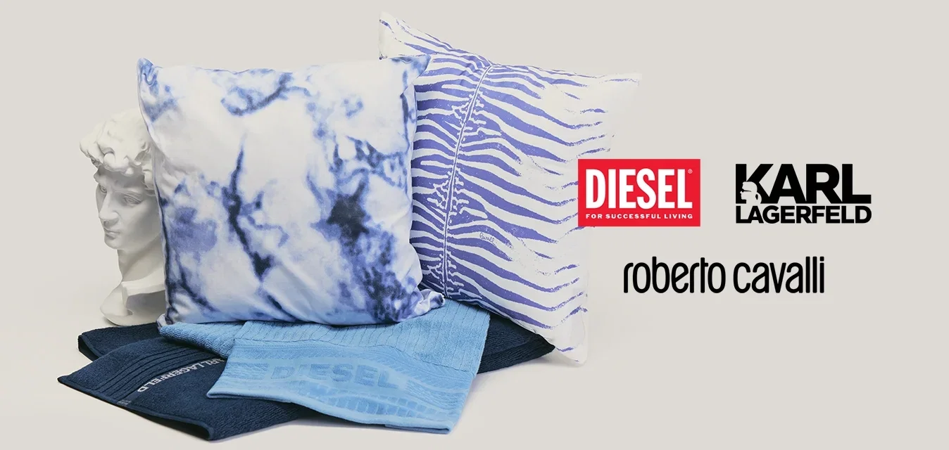 Dos almohadas decorativas con patrones azules, dos tejidos azules doblados y un busto blanco de estilo clásico sobre un fondo neutro.