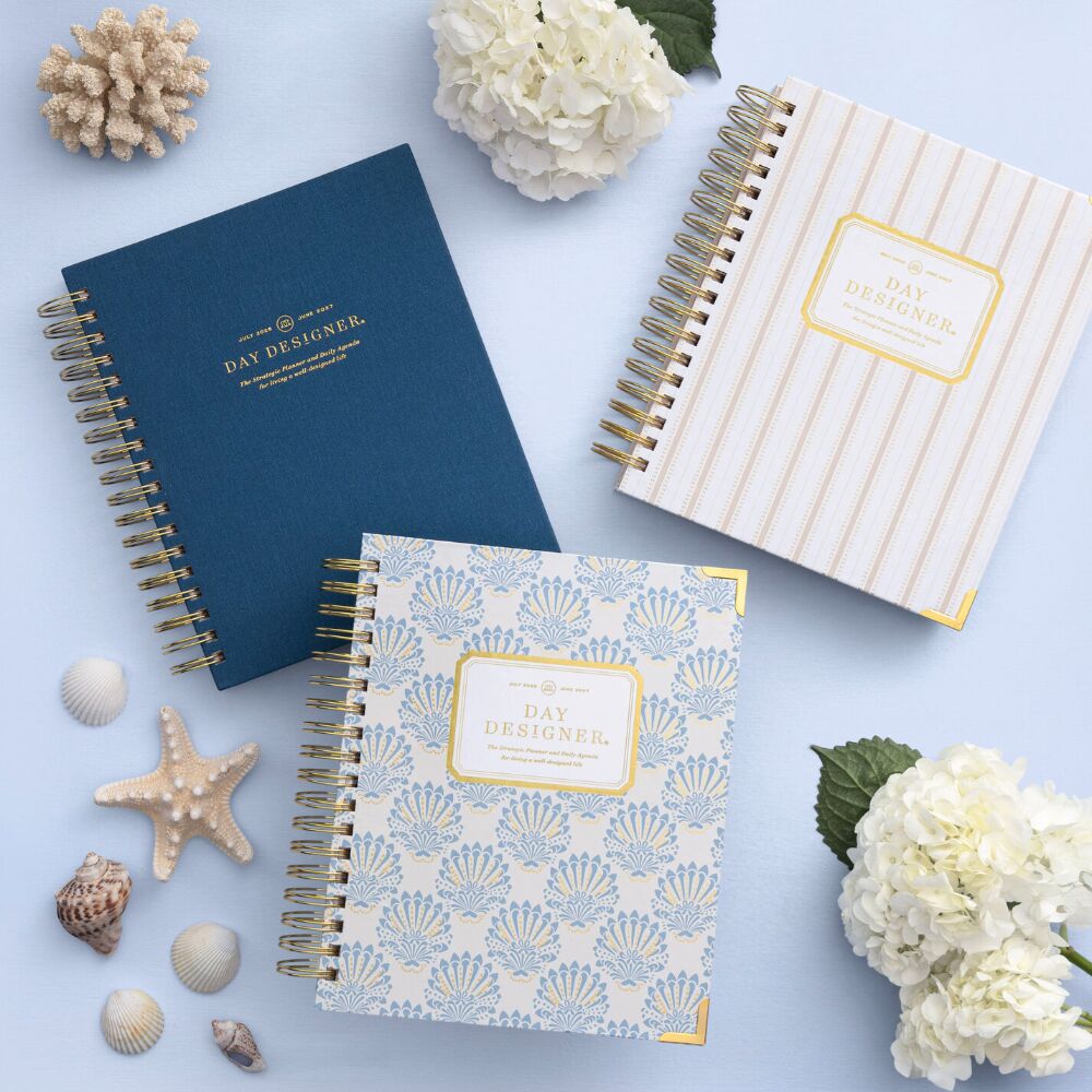 stacked day designer planners in navy blue, gold striped, and repeated blue shell pattern, white flowers, sea shells, light blue background