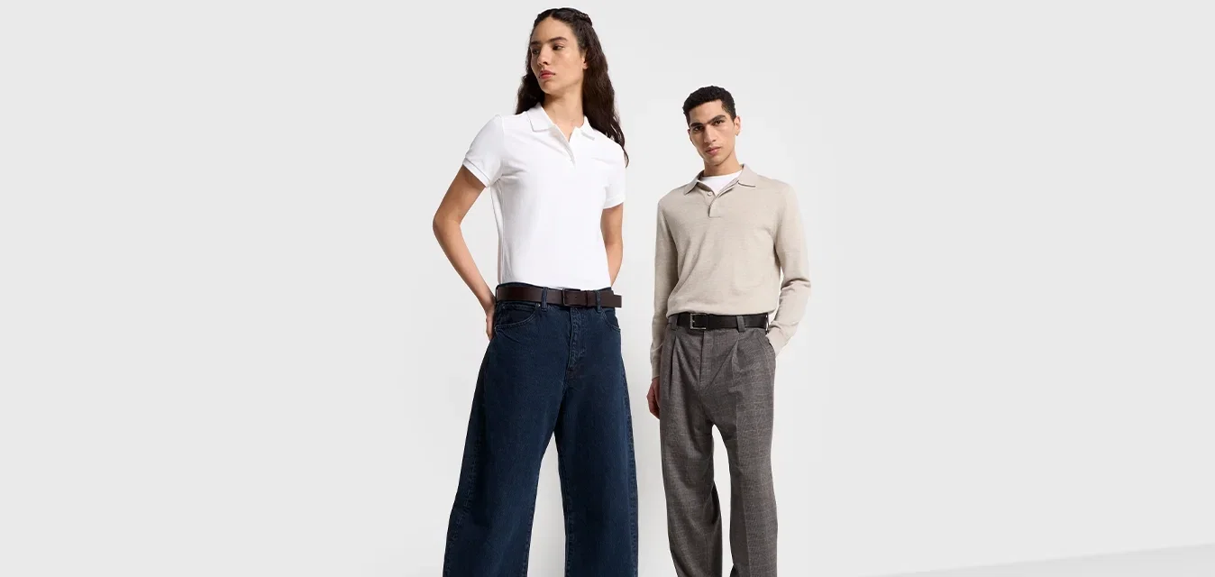 Twee modellen staan tegen een lichtgrijze achtergrond. De ene draagt een witte polo en donkere wijde broek; de ander een beige trui en grijze broek.