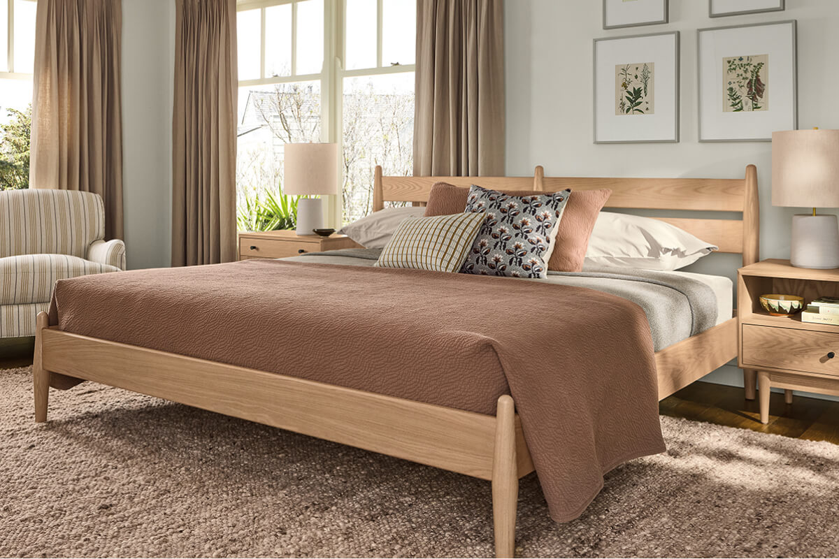 Grove bed with light wood frame, brown bedding, and matching nightstand beside windows with curtains.