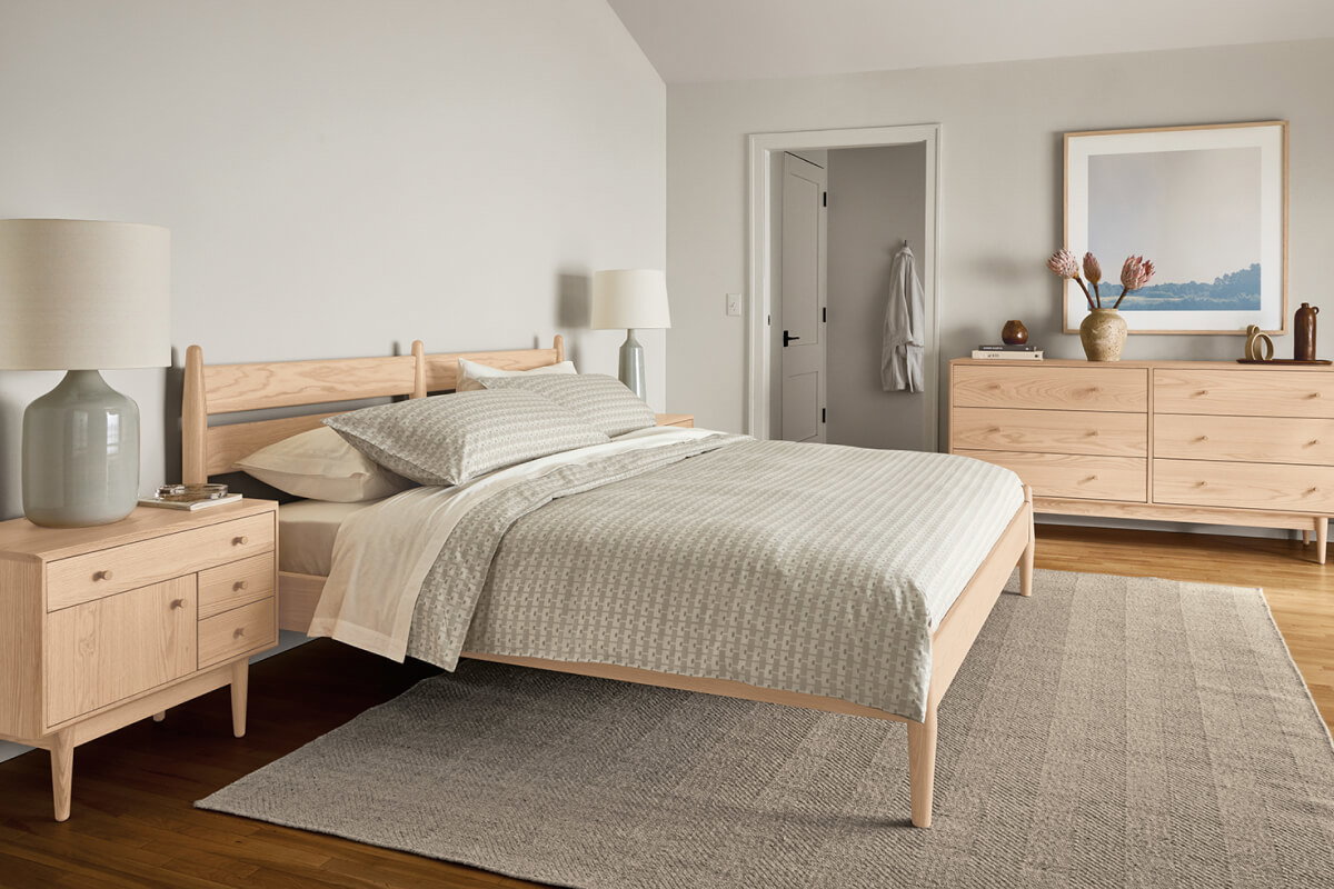 Grove bed in light wood with grey bedding, surrounded by matching nightstands and dresser with table lamp and decor.