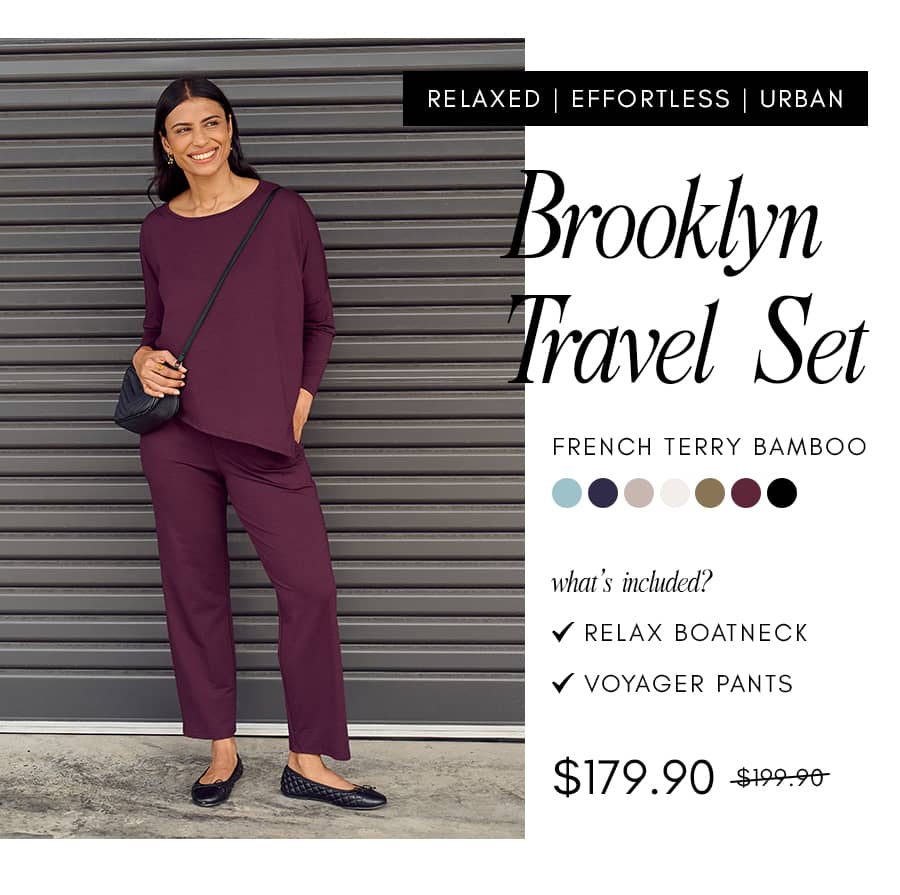 A woman stands in front of a grey corrugated metal wall wearing a plum-colored long-sleeved top and matching pants, holding a black quilted purse, wit...