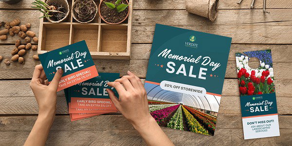 Two hands arrange Memorial Day sale postcards on a wooden table alongside a matching flyer and door hanger. Potted plants line the back of the table in a wooden organizer.