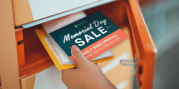 A hand places Memorial Day sale postcards into an orange mailbox slot.