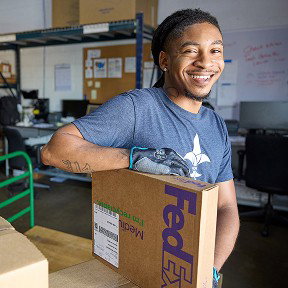 A smiling FedEx employee rests his arm on a package in a warehouse setting.