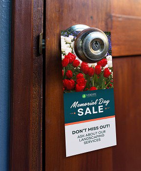 A Memorial Day sale door hanger featuring red and white flowers hangs on the knob of a wooden door.