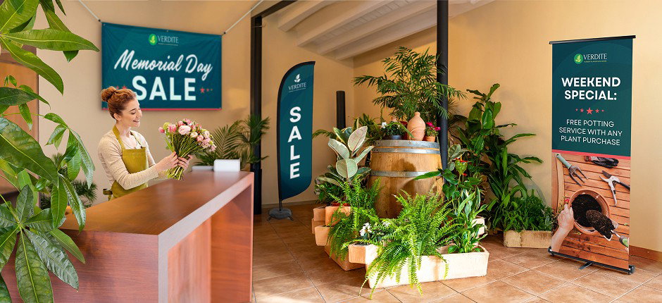 A plant store interior features a woman holding a bouquet at the counter, surrounded by plant displays. A Memorial Day sale sign hangs in the background next to a matching flutter flag. A large retractable banner promotes free potting services.