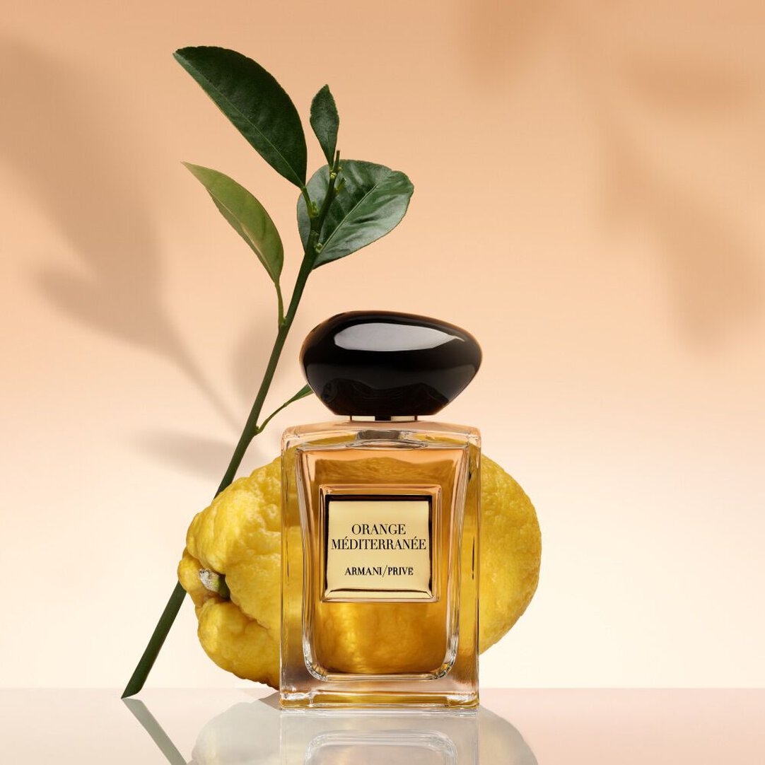 Vea un frasco de Armani/Privé Orange Méditerranée presentado junto a una naranja amarga fresca y hojas verdes sobre un fondo soleggiato.