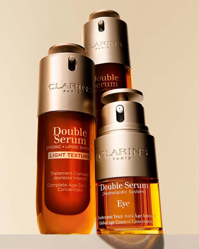 Three bottles of Clarins Paris Double Serum on display, including Light Texture and Eye versions, with a golden backdrop.