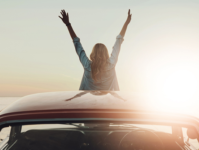 IMAGE OF A WOMAN' SITTING ON THE BONNET OF A CAR WATCHING THE SUNSET WITH HER HANDS UP IN THE AIR