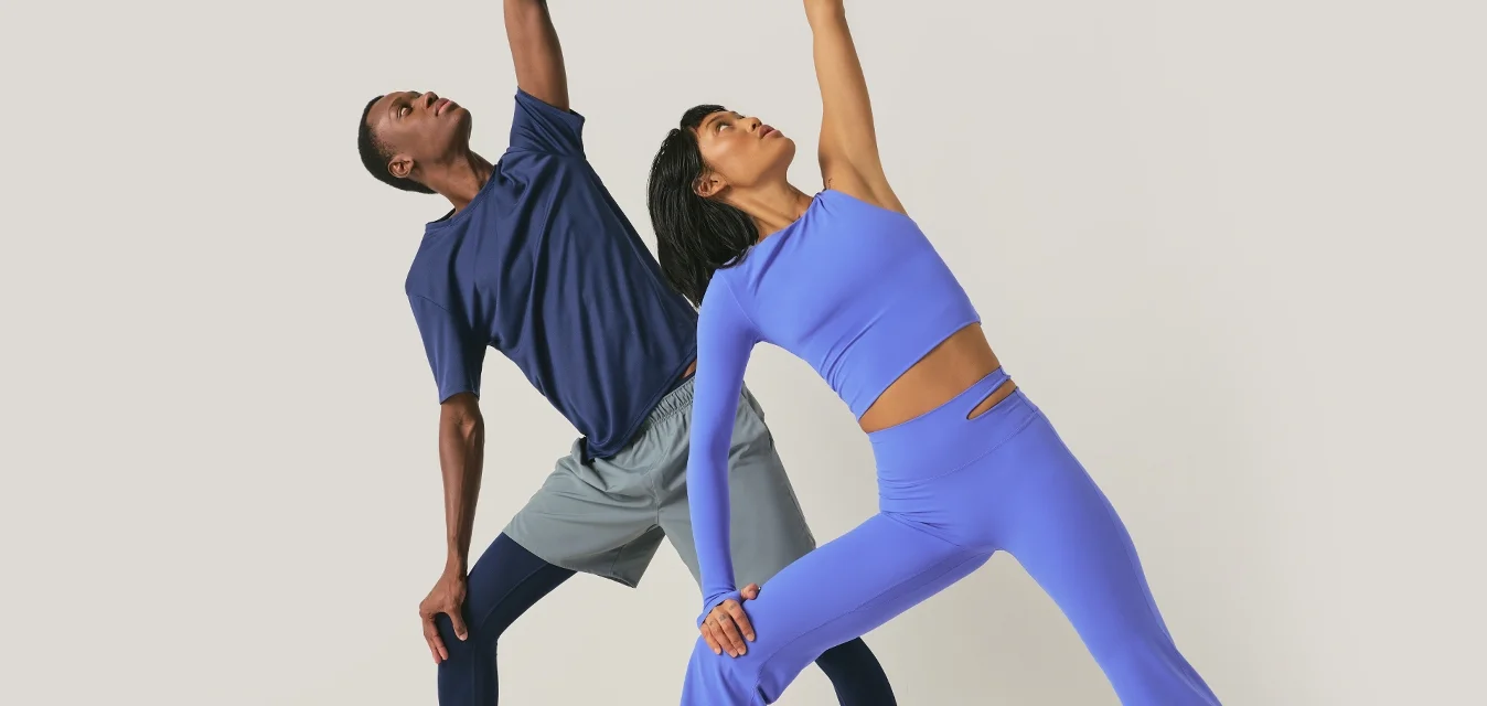 Twee mensen in sportkleding die een yoga-zijstretch uitvoeren, met één arm omhoog reikend en de andere arm die hun been aanraakt.
