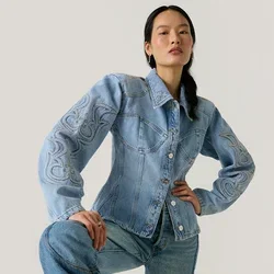 Vrouw met lang donker haar die een lichtblauwe geborduurde spijkerjas en bijpassende jeans draagt, poserend met één knie omhoog tegen een effen achtergrond.