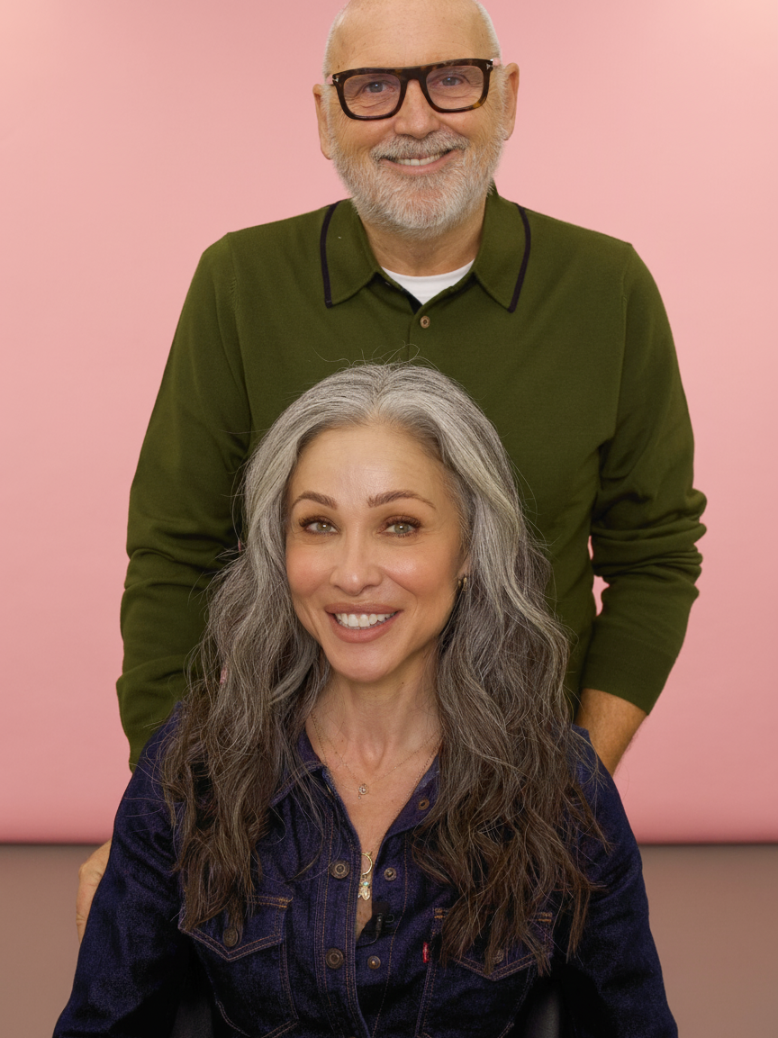 A smiling woman with gray hair in a denim shirt is in front of a man wearing glasses and a green sweater.