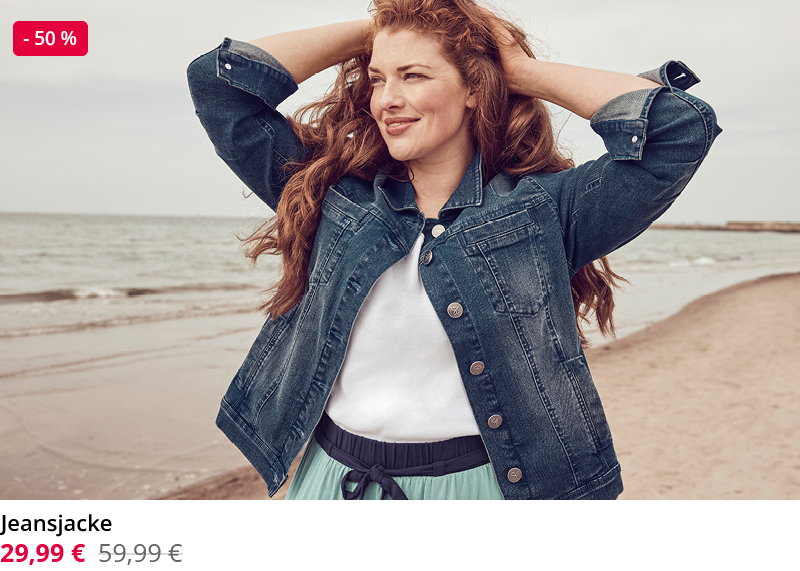 Frau mit lockigen roten Haaren posiert lächelnd am Meer und trägt eine blaue Jeansjacke über einem weißen Shirt