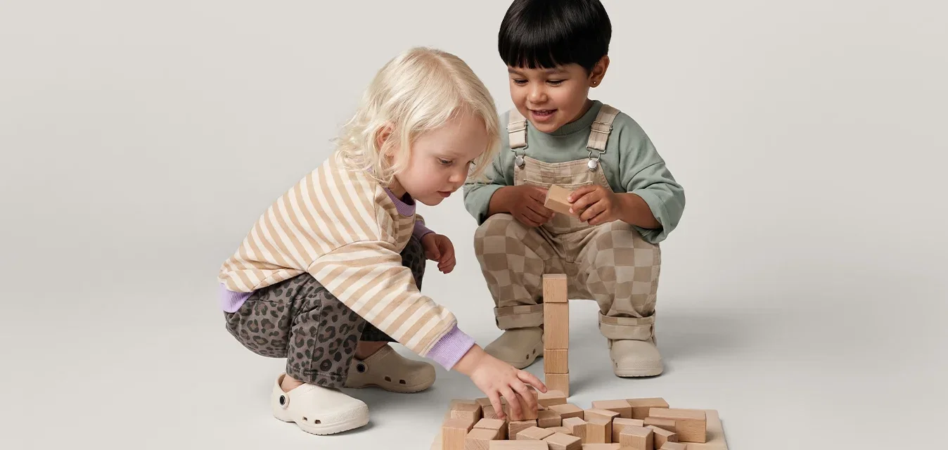 Dos niños pequeños sentados en el suelo, jugando con bloques de madera dispuestos sobre una tabla plana con un fondo liso.