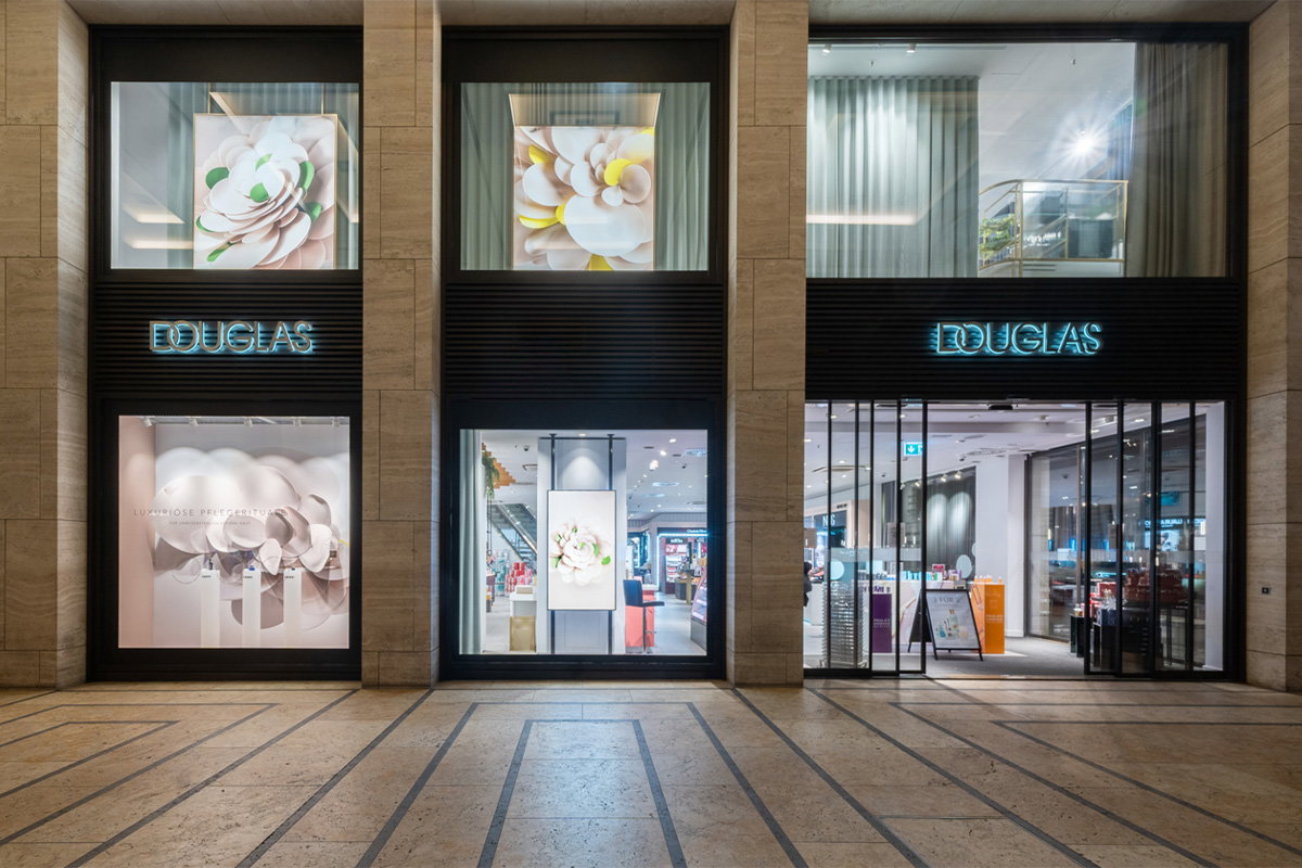 Fassade eines modernen, gut beleuchteten Douglas-Geschäfts mit zwei großen Schaufenstern mit floralen Motiven und Glastüren, die einen hell erleuchteten Innenraum zeigen, der Eleganz ausstrahlt.