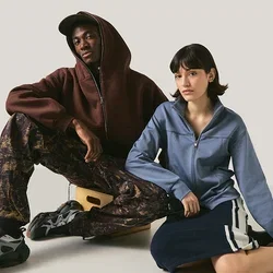 Twee mensen zitten op een eenvoudige vloer tegen een neutrale achtergrond, de een draagt een hoodie en met patroon bedrukte broek, de ander een blauwe jas en rok.