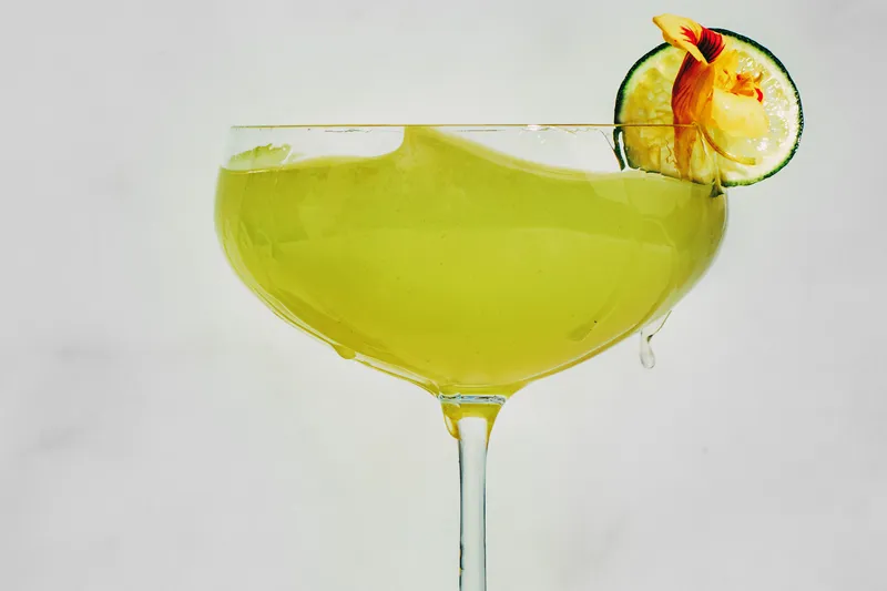 A chartreuse-colored cocktail in a coupe, garnished with a lime wheel and an orchid.