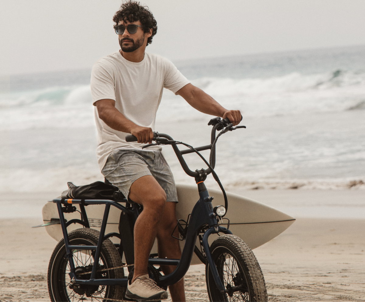 Man riding a bike on the beach with a surfboard, wearing casual coastal outfit under headline “The all-day explorer.”