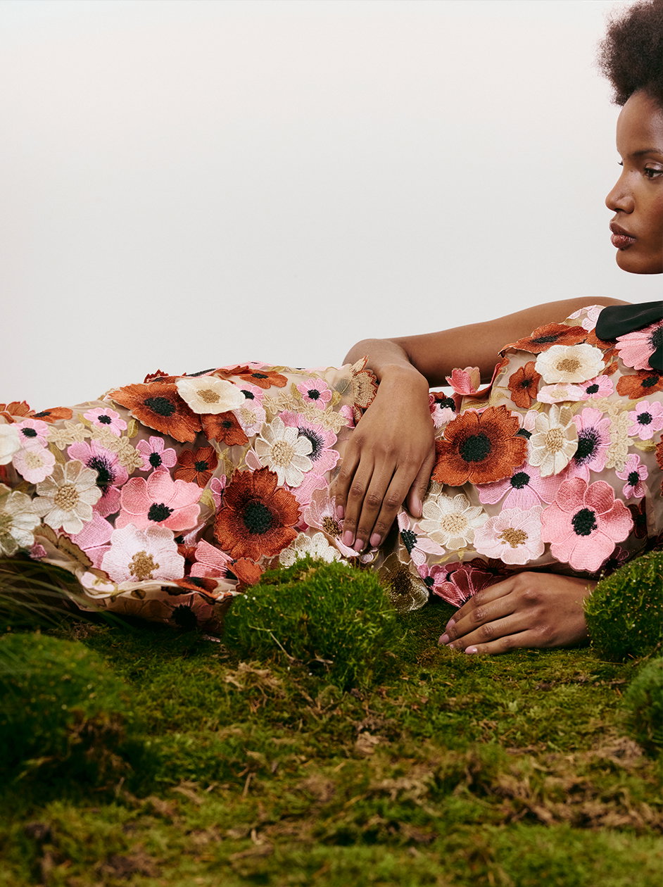 Ein Model von DOROTHEE SCHUMACHER ruht auf weichem grünem Moos. Sie präsentiert ein kunstvolles Kleid mit opulenten 3D-Blütenapplikationen in Nuancen von Rosa, Rostorange und Creme – ein Look, der feminine Eleganz und florale Inspiration vereint.
