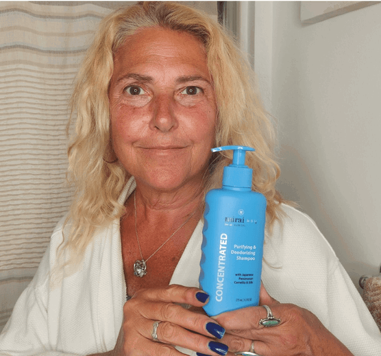 A woman with blonde hair, wearing a white robe and a necklace, holds a blue bottle of (square)