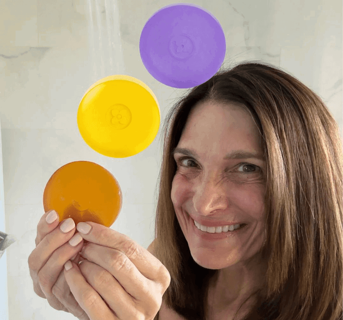 A smiling woman with brown hair holds up an orange-colored disc in her hands, with a yellow disc and a purple disc floating above her shoulder in a shower. (square)