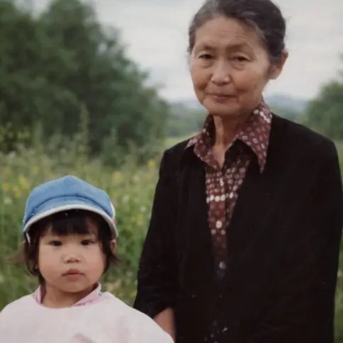 An elderly Asian woman and a young Asian girl stand side-by-side in a grassy field, with trees in the background. (square)