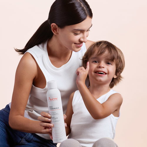 Smiling mother and young child in white tops holding Avène Thermal Spring Water spray bottle, gentle skincare moment
