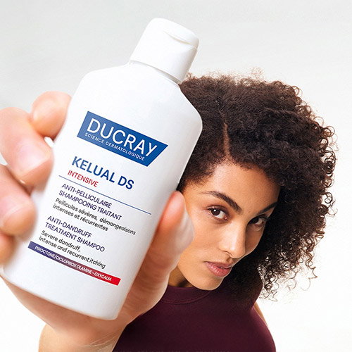 Young woman with curly hair holding a Ducray Kelual DS Intensive anti-dandruff treatment shampoo bottle