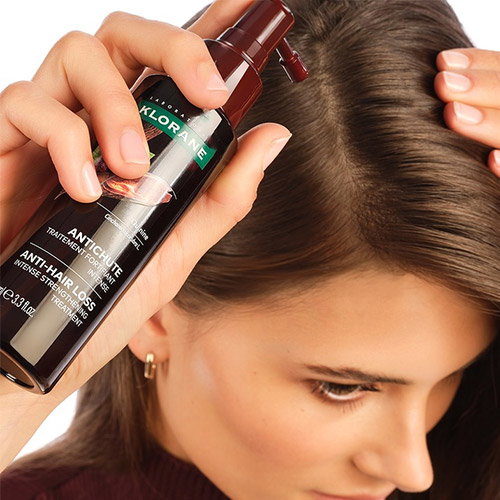 Close-up of a woman applying Klorane anti-hair-loss scalp treatment spray to a thinning hair parting