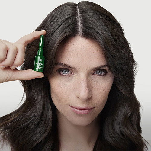 Woman with long dark hair and freckles holding a small green hair serum vial beside her temple against a neutral background
