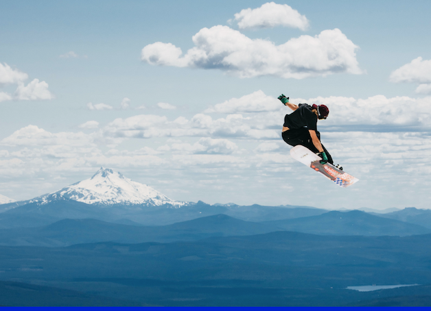 Snowboarder in the air with snowy mountains and a cloudy sky in the background