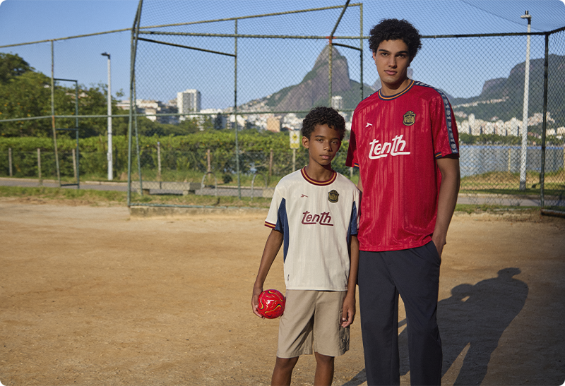 Two boys in football jerseys, one holding a red ball, standing on an urban dirt pitch with a fenced backdrop and distant city skyline