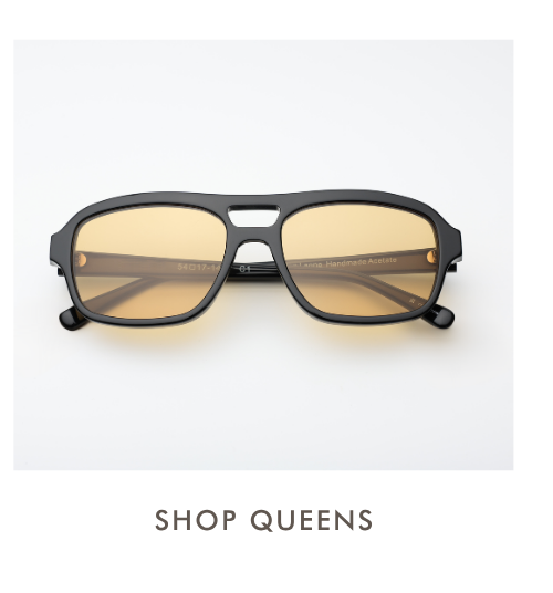 A studio product shot of the Queens sunglasses. These feature a black acetate frame with a squared-off aviator shape, a double bridge with a rectangular cutout, and yellow-tinted lenses. Text below reads: 