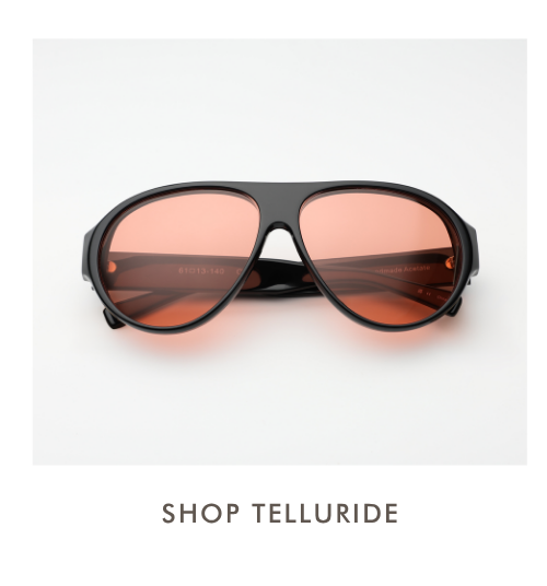 A studio product shot of the Telluride sunglasses. The frames feature a bold, oversized black acetate aviator silhouette with a continuous brow line and deep orange-tinted lenses. Text below reads: 