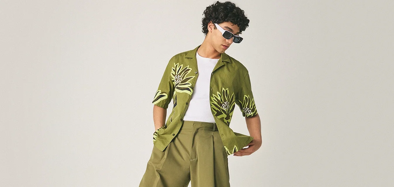 Jongere in olijfgroene wijde broek, gestreepte groene shirt, witte top, zwarte sandalen en zonnebril, poserende op een lichte achtergrond.